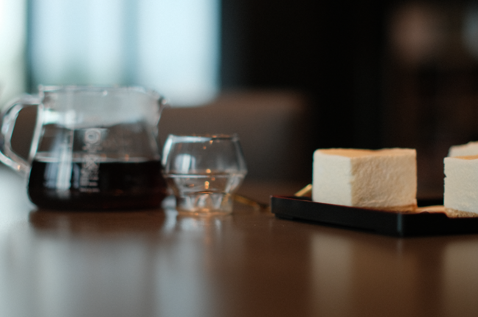 A glass coffee carafe and a small glass cup sit on a wooden table beside a tray with two square desserts. The setting feels calm and inviting.