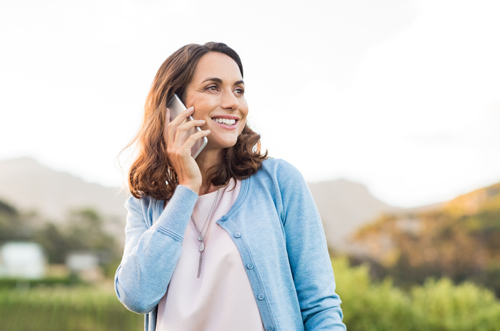 Dark haired woman in a pale blue cardigan standing outside talking on a cellphone.  