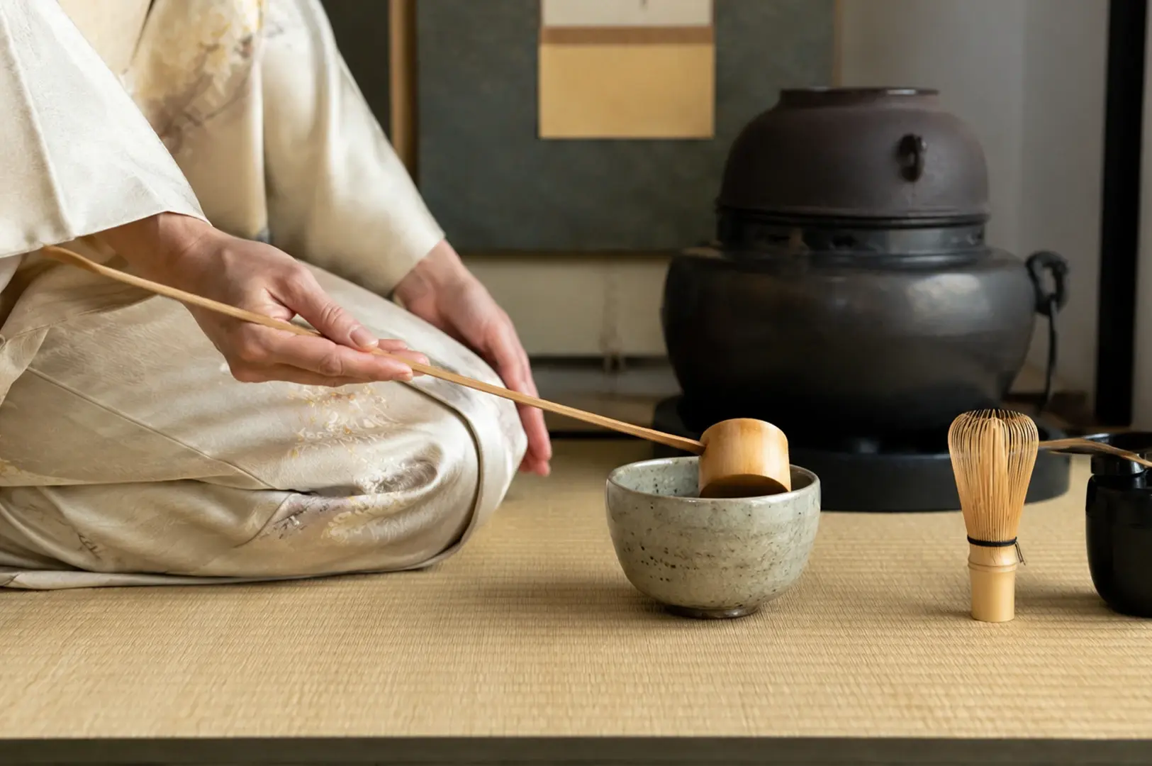 A close-up photograph focusing on the elegant hands of a practitioner dressed in a cream-colored, patterned kimono kneeling on a tatami mat. The person is holding a long, slender bamboo ladle, carefully preparing to scoop water into a light gray, speckled ceramic tea bowl positioned in front of them. To the right, a bamboo tea whisk stands upright next to a black ceramic vessel, while a large, dark iron kettle on a black stand sits in the background against a muted screen.