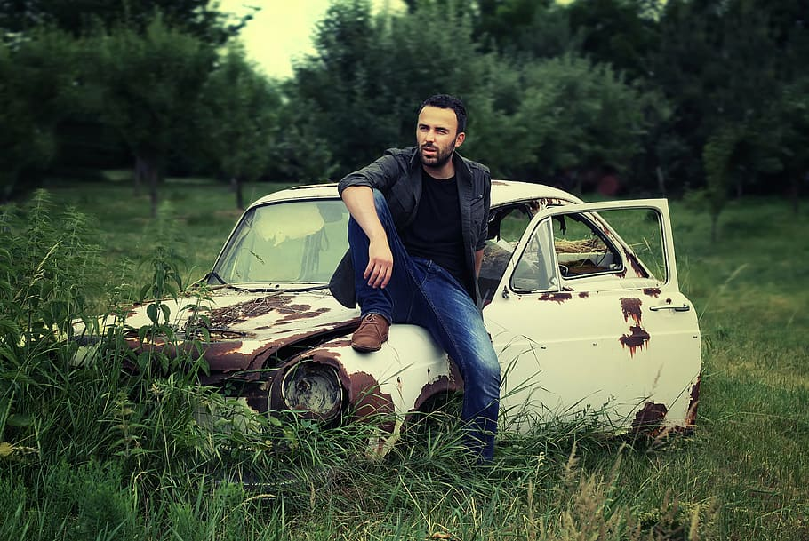 A man standing in front of a junk car, smiling, with a stack of cash in his hand