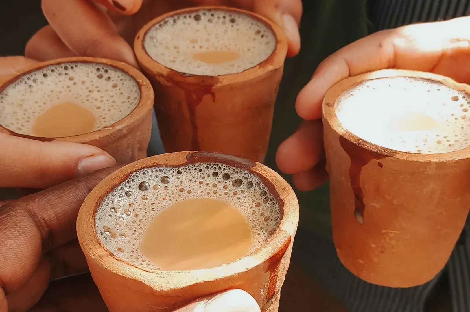 A group of people hold up small, traditional unglazed clay cups filled with hot, steaming masala chai, with the focus on the foam and the warm brown liquid inside, capturing a communal moment of sharing a beverage.