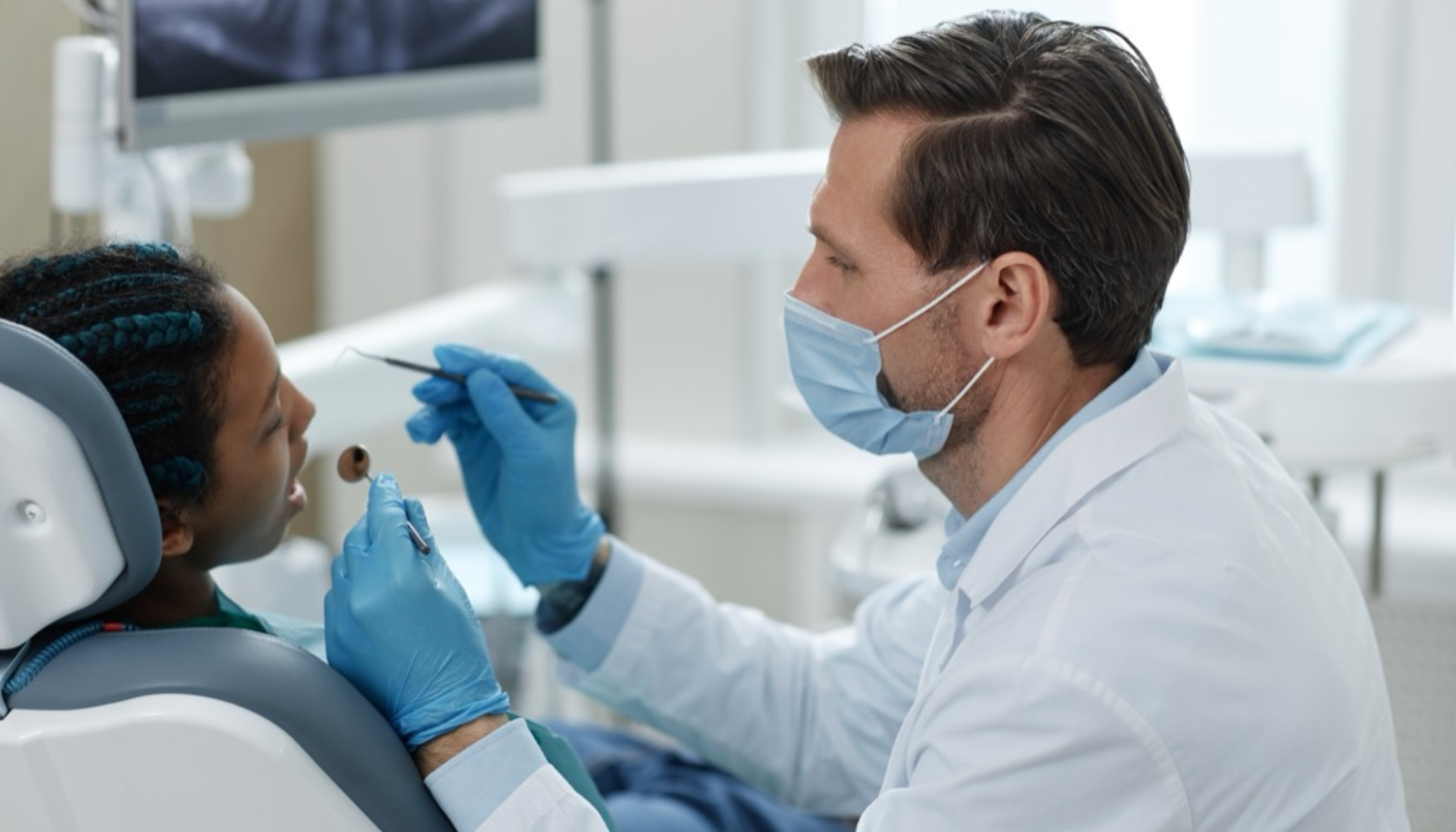 Tannlege med munnbind undersøker munnen til et barn i tannlegestol, med instrumenter i hånden i moderne klinikkmiljø.