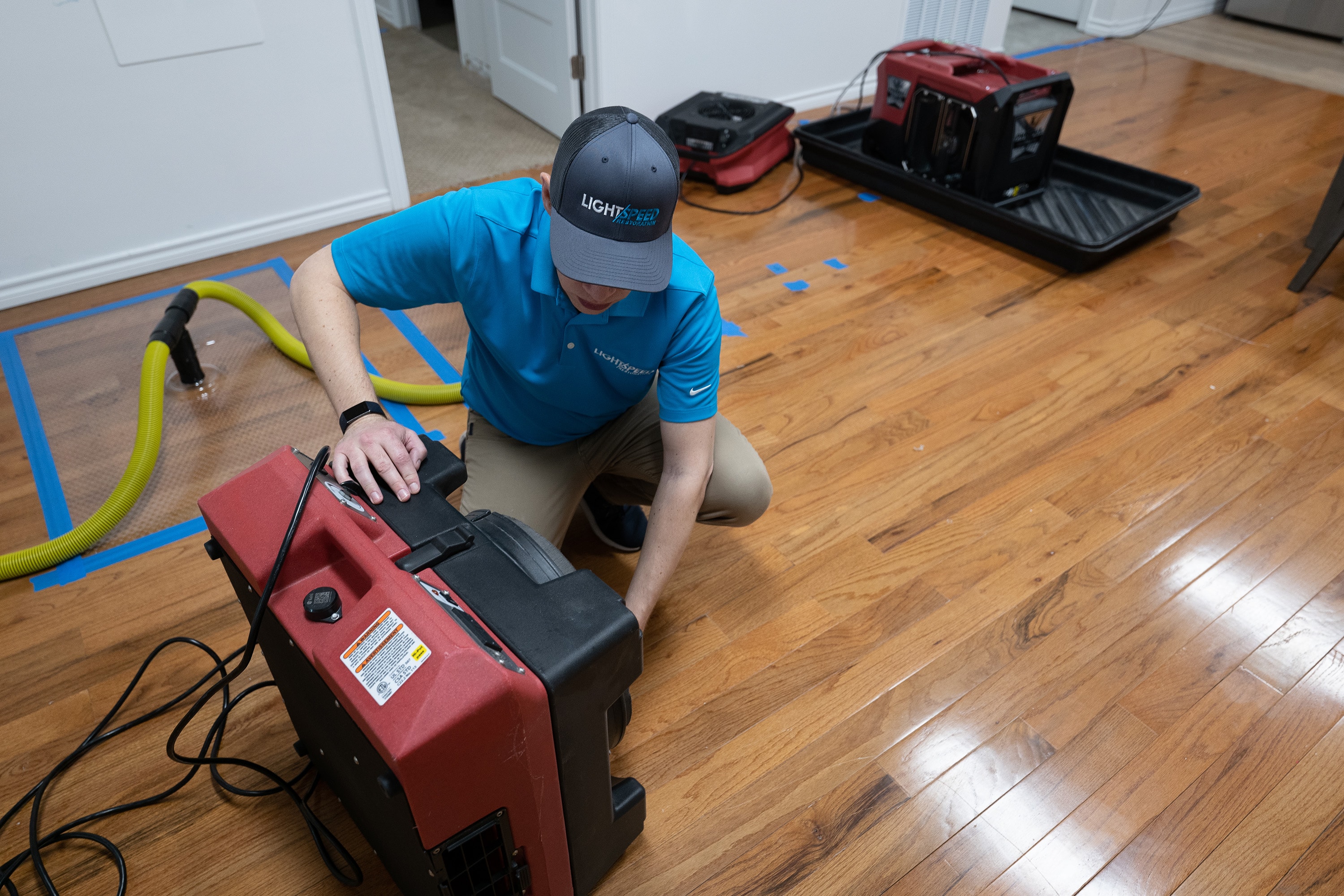 The image shows multiple industrial dehumidifiers and air movers set up in a living room with an exposed subfloor, indicating a flood damage restoration process. The equipment is positioned to remove moisture and prevent mold growth from the damp environment created by flood water.