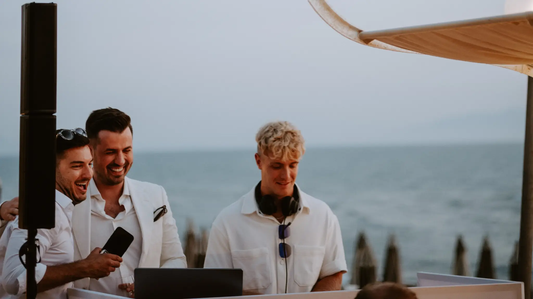 DJ Nathan White mit Kopfhörern steht an einem Pult am Meer, während zwei elegant gekleidete Männer daneben lachen und ein Mikrofon halten; im Hintergrund sind Strand, Sonnenschirme und das offene Meer zu sehen.
