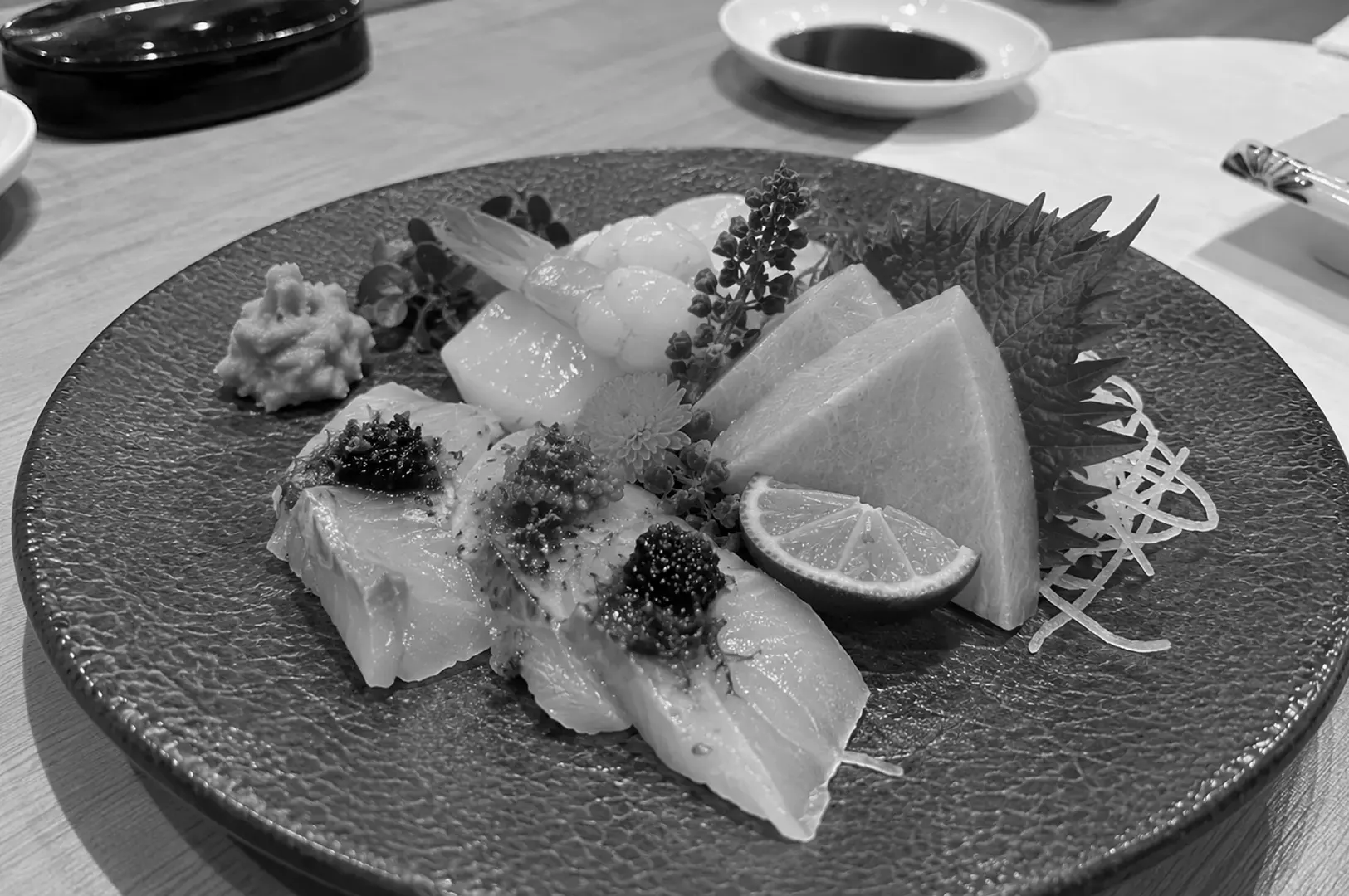 Large textured plate of assorted sashimi served with grated daikon, shiso leaves, edible flowers, wasabi, and a lime wedge.
