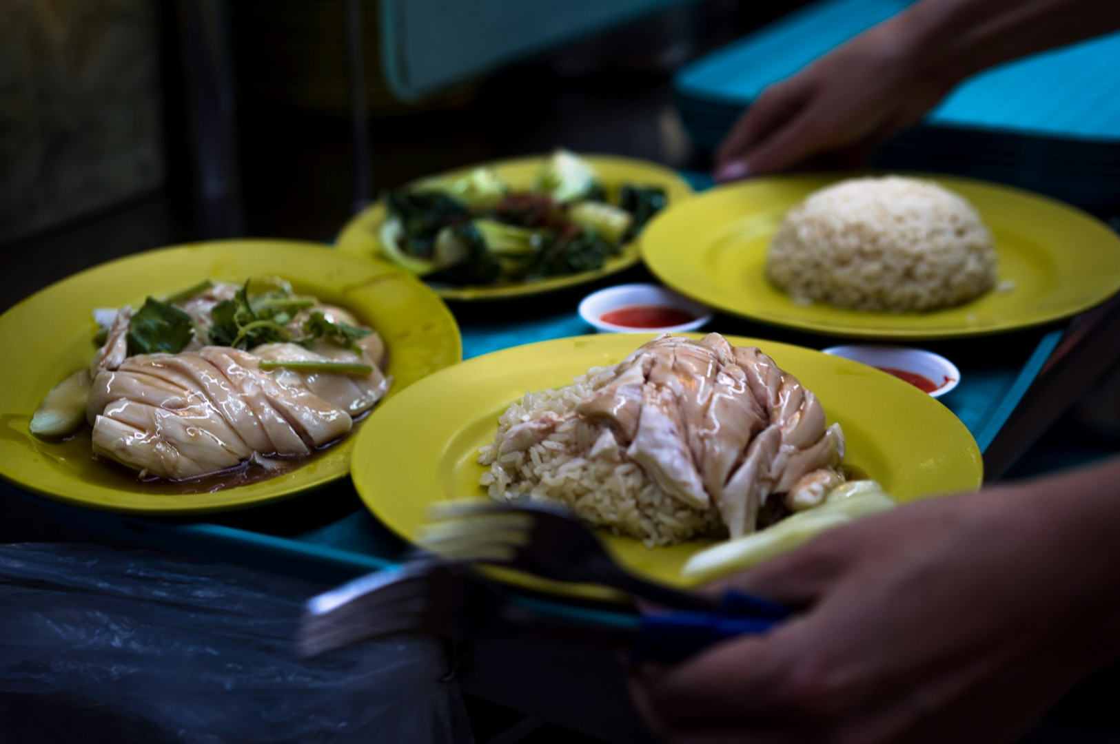 Plates of Hainanese chicken rice with steamed chicken and white rice on yellow plates, garnished with sauces. A hand serves a dish, creating a cozy street food ambiance.