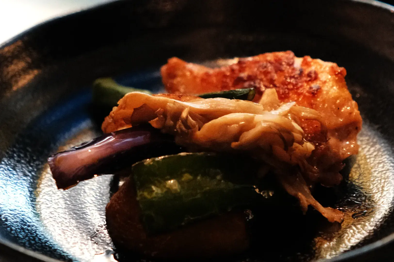 A plate of food featuring a piece of chicken with sautéed vegetables, including green peppers and a mushroom, served on a dark plate.