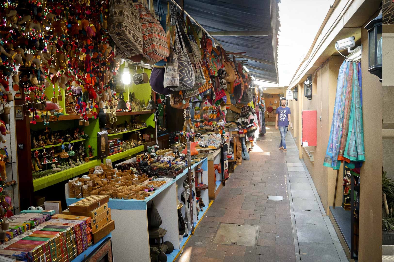 A narrow corridor in Little India Arcade lined with colorful stalls selling handicrafts, bags, ornaments, and textiles.