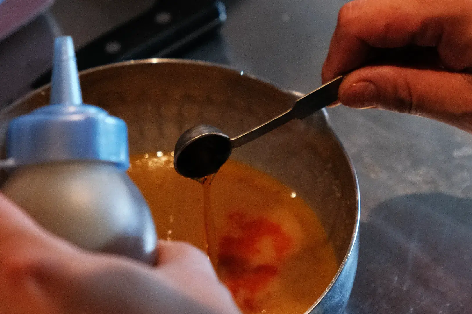 A person's hand pouring a liquid ingredient from a squeeze bottle into a metal mixing bowl that contains a mixture of orange and yellow liquid, with a measuring spoon also seen in the scene.