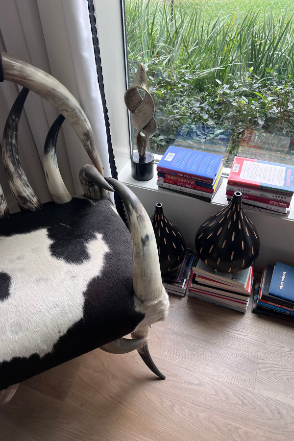 Cowhide accent chair by a window with stacked books, sculptural vases, and tall greenery outside.