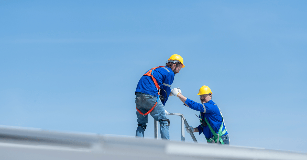 Two roofers wearing safety gear, working together on a commercial roofing project under a clear blue sky.
