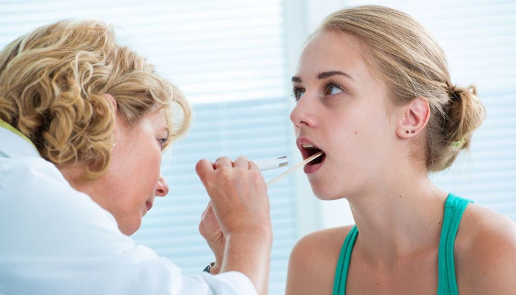 Lege undersøker halsen til ung kvinne med lommelykt og trepinne under en medisinsk konsultasjon på legekontor.