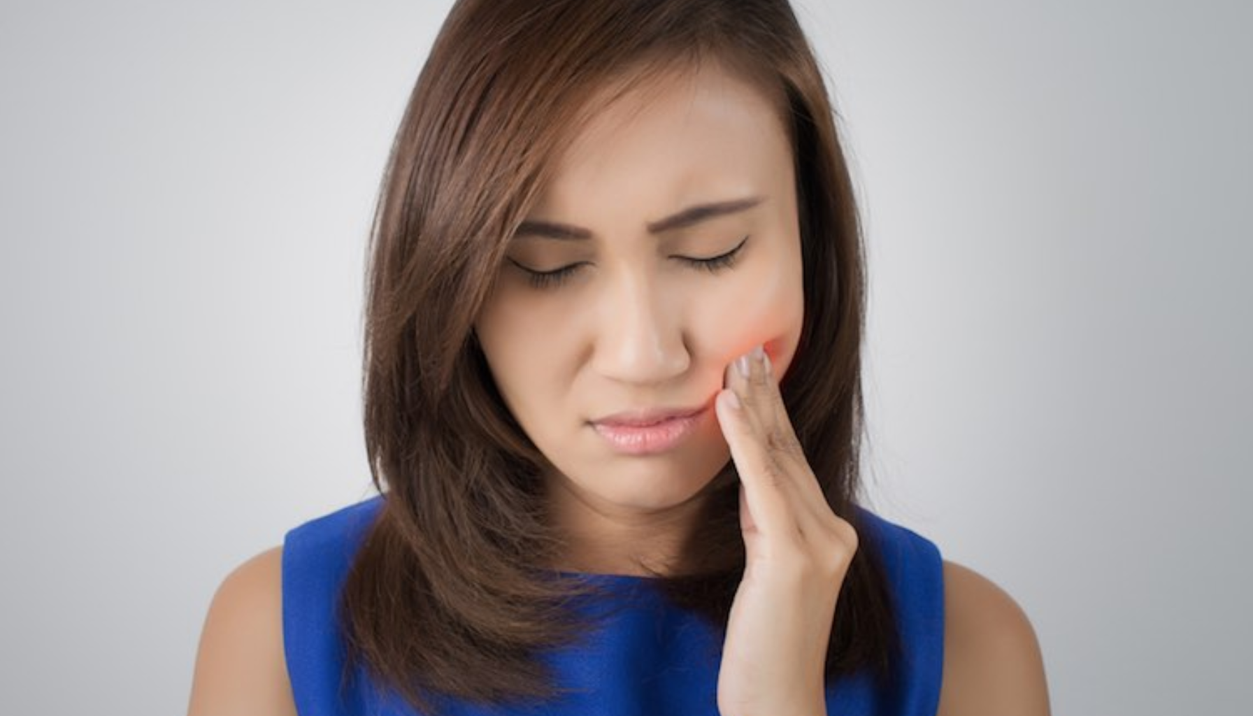 Kvinne med blå topp holder hånden på kinnet og uttrykker smerte, antyder tannverk eller kjevesmerter.