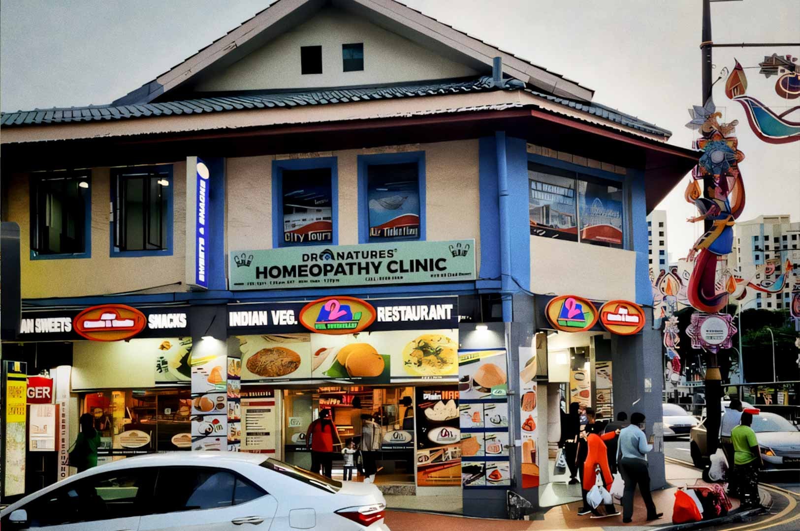 A corner shophouse on Syed Alwi Road featuring an Indian vegetarian restaurant on the ground floor and a homeopathy clinic above, with people walking past and cars on the street.