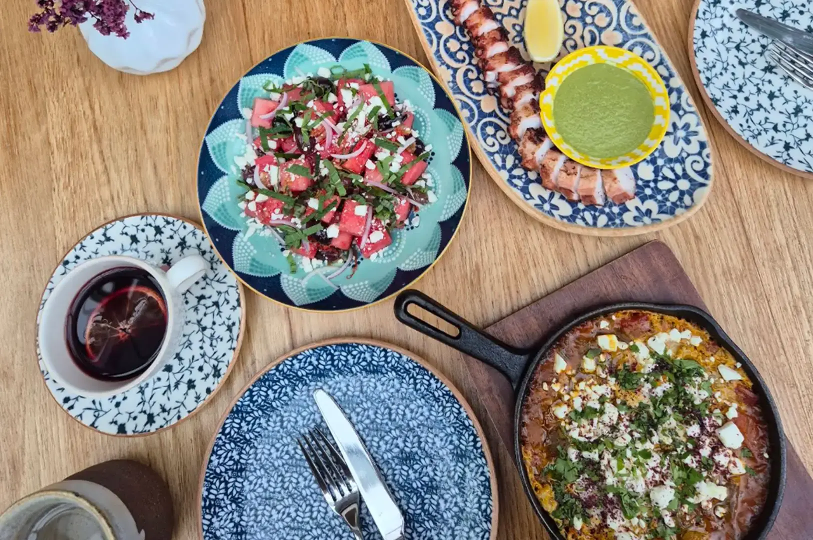 A flat-lay overhead view of a wooden dining table arranged with a cast-iron skillet dish topped with herbs and cheese, a colorful watermelon and feta salad on a patterned plate, a grilled octopus tentacle with green dip, cutlery, and a hot cup of tea.