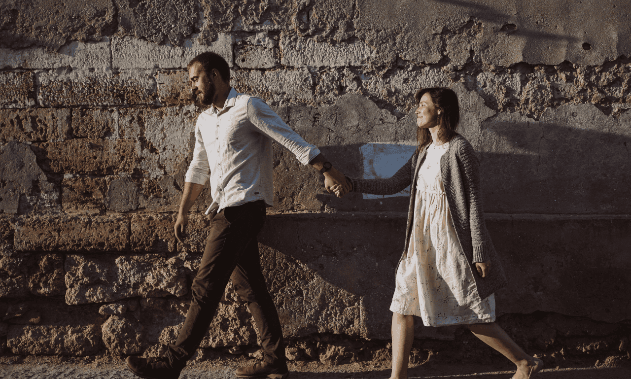 A guy and girl walking holding hands with a brick wall behind them.
