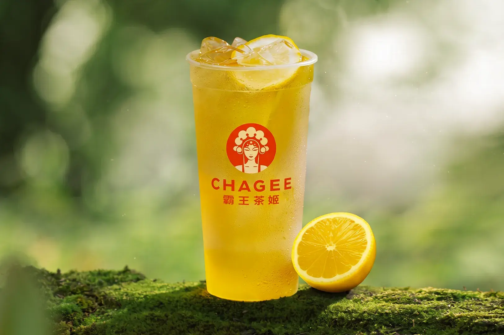 Branded CHAGEE iced lemon tea cup resting on a mossy surface with a fresh lemon cut in half beside it.