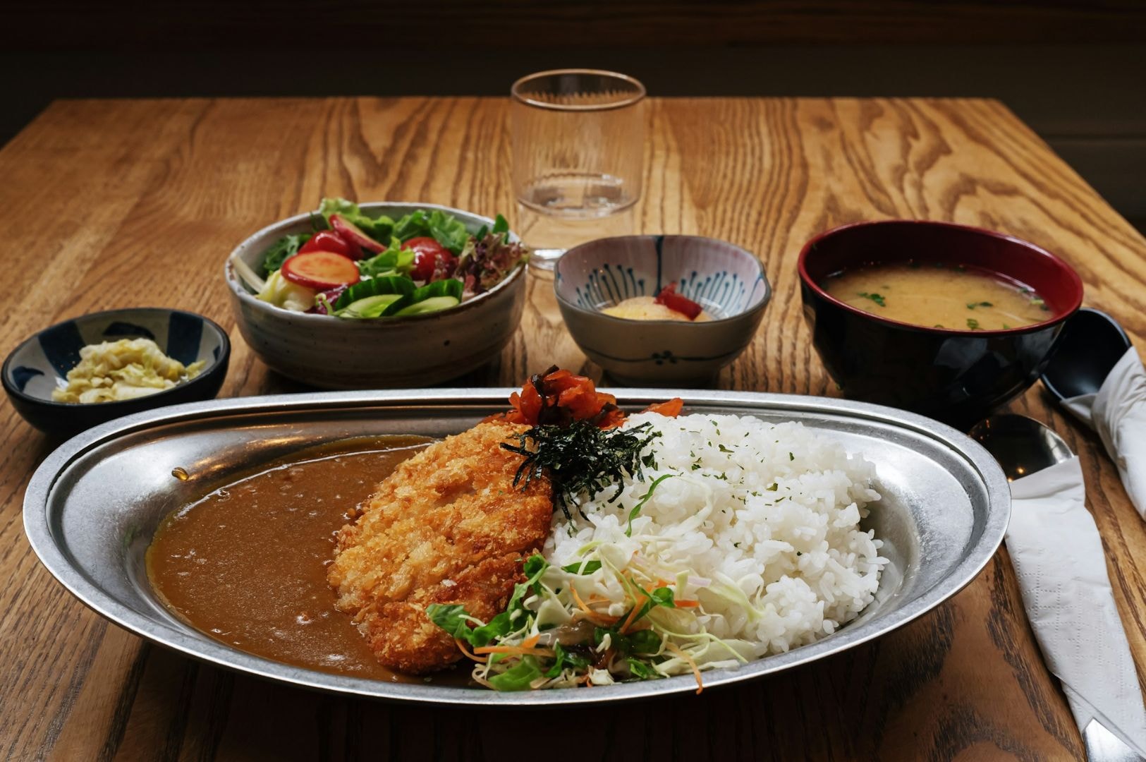 Plate of curry rice with breaded cutlet, garnished with vegetables and seaweed on a wooden table. Accompanied by salad, soup, and side dishes.