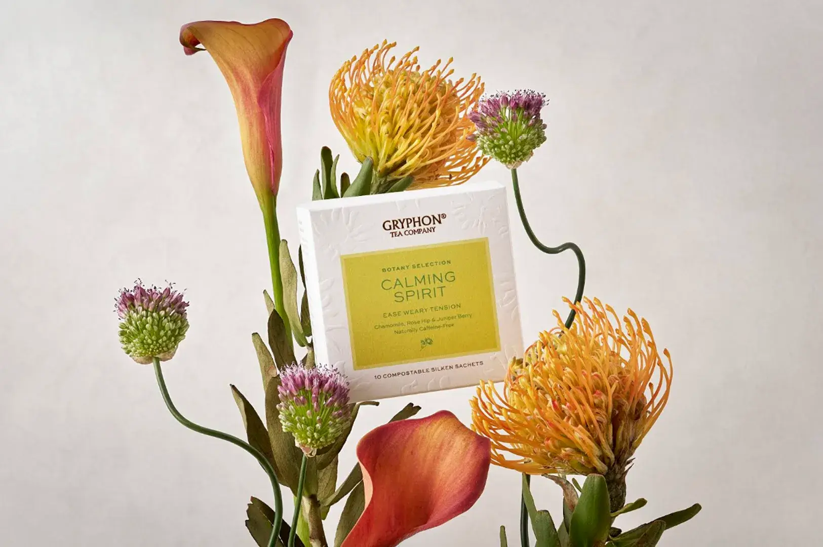 A close-up shot of the white box packaging for Gryphon Tea Company's "Calming Spirit" tea from their Botany Selection, nestled artistically amid a striking floral arrangement against a neutral beige background. The tea box features a lime-green central label detailing the blend as caffeine-free with chamomile, rose hip, and juniper berry. It is framed by a dynamic cluster of unique botanical stems, including two vibrant, orange-tipped yellow pincushion proteas, a pair of tall, elegant calla lilies in warm sunset shades of orange and pink, and several slender green stalks sporting round, spiky purple and green allium flower buds.