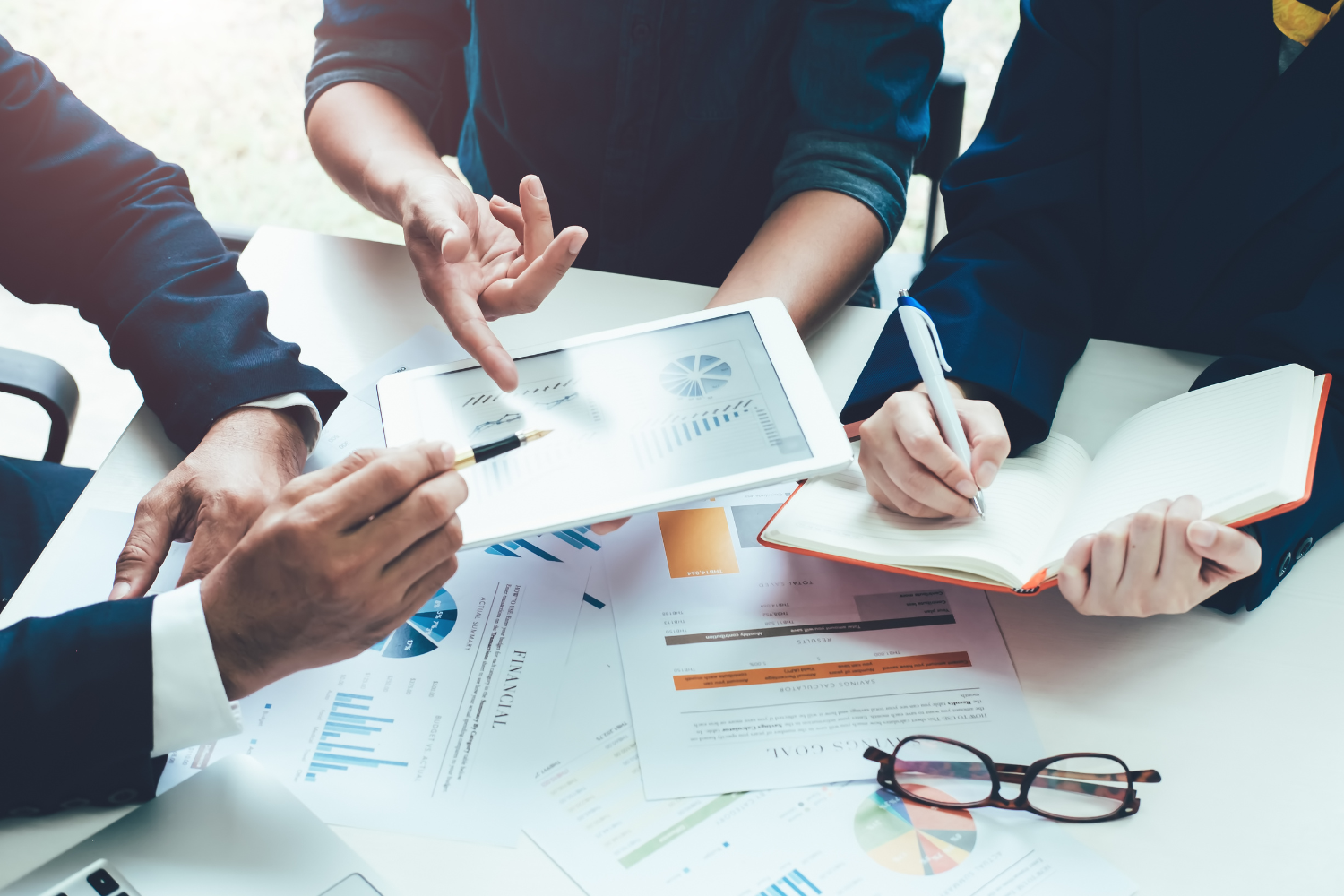 A meeting with financial charts, glasses, tablet and notebook on a white table.