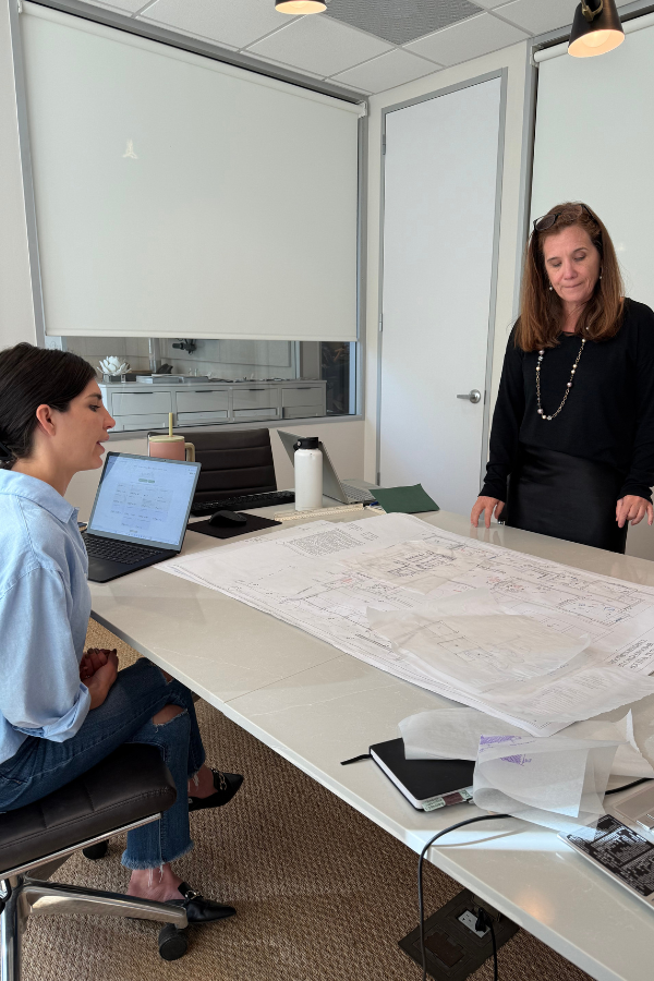 Two women discuss architectural plans laid out on a large white conference table. One is seated with a laptop open, while the other stands, reviewing the drawings.