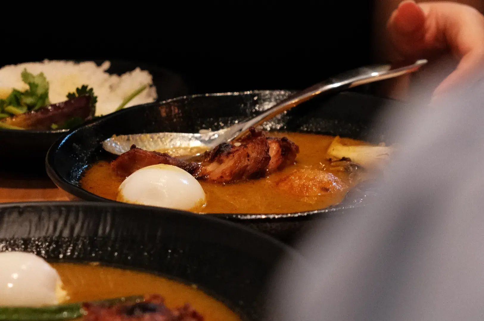 A hand lifts a spoon from a black bowl of rich orange soup with a soft-boiled egg and browned meat, creating a warm, hearty dining scene.