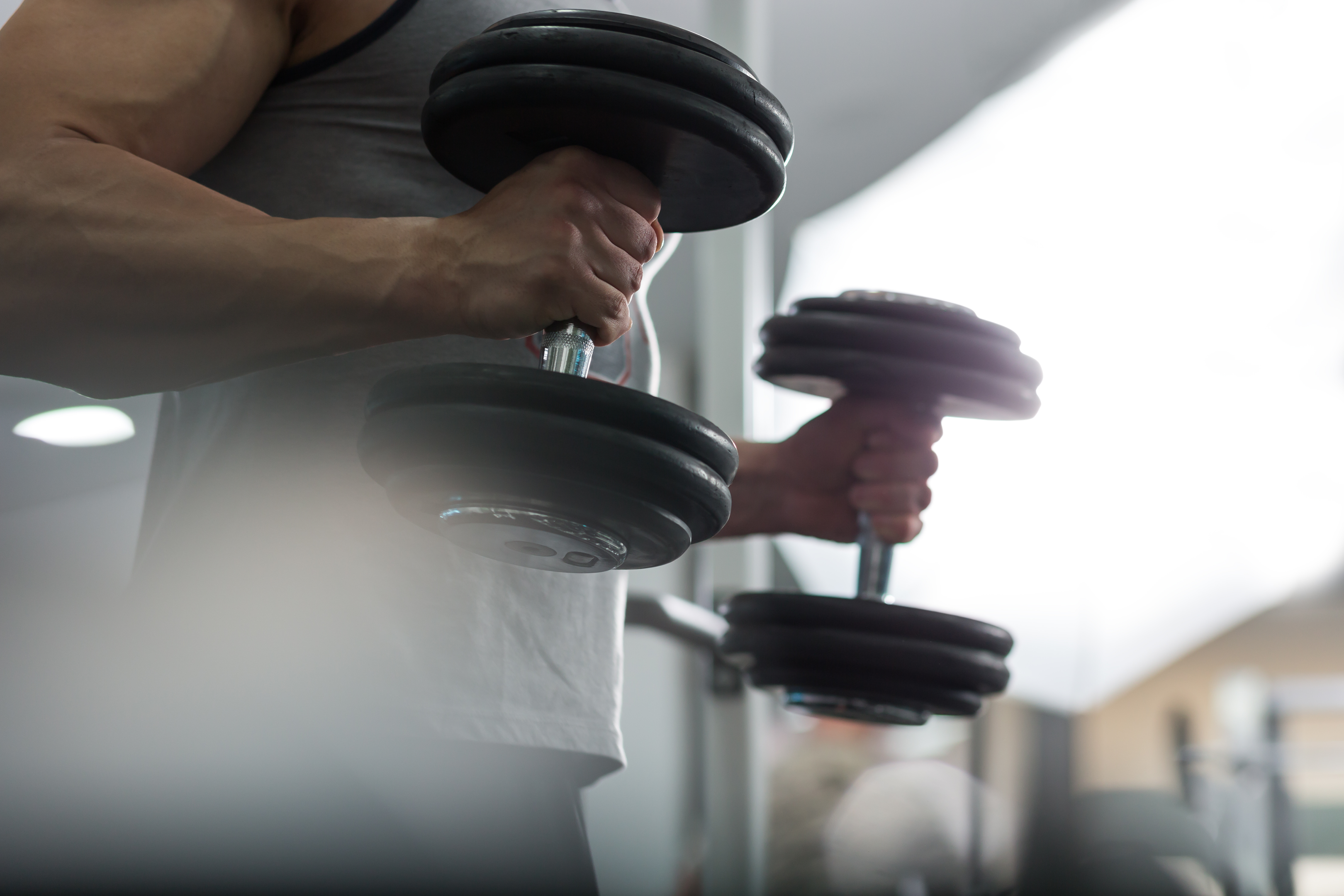 Shot of midsection of a white male with visible bicep muscles, facing right and holding large dumbbells in each hand