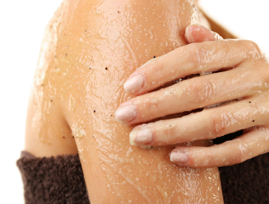 Een persoon brengt een grove bodyscrub aan op de schouder, waarbij de hand zichtbaar over de scrub wrijft om de huid te exfoliëren. Dit gebeurt  vaak in roterende bewegingen.