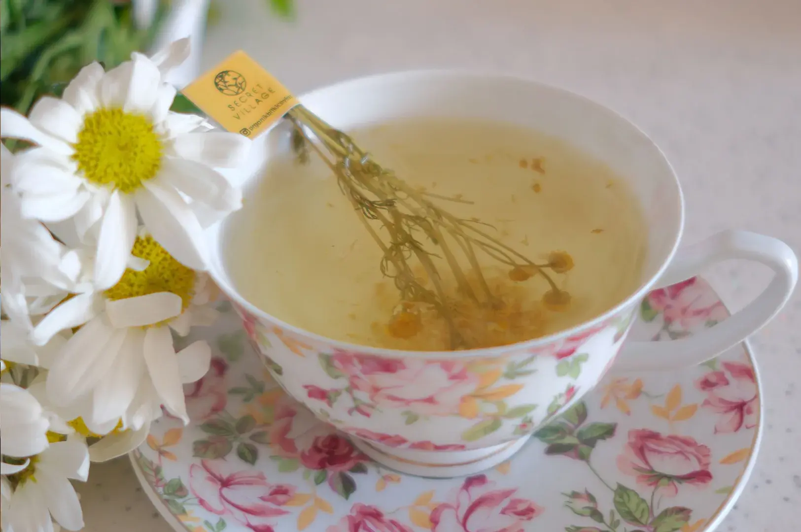 A close-up shot features an elegant white ceramic teacup and saucer adorned with a vibrant pink and green floral rose pattern, filled with a pale yellow chamomile infusion. Submerged in the tea is a bundle of whole dried chamomile stems with a yellow tag from "Secret Village" resting on the rim. Fresh white daisies with bright yellow centers lean against the left side of the cup, set against a soft, bright background that enhances the delicate, refreshing feel of the scene.