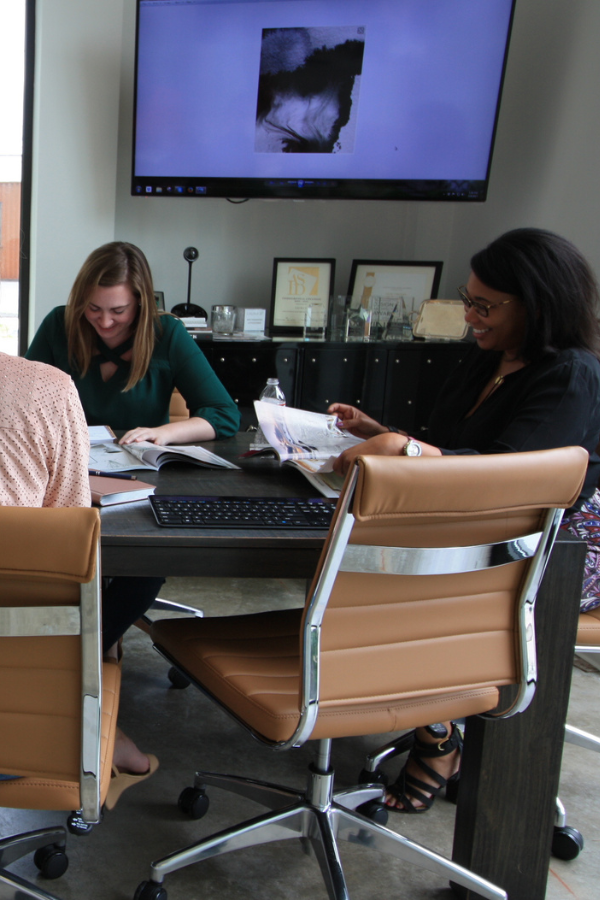 A small group of designers sit around a conference table reviewing catalogs and material books, with a large screen displaying artwork on the wall behind them.