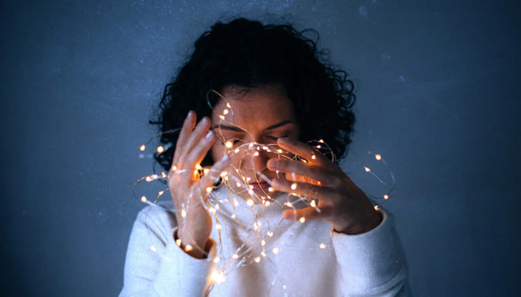 Trigeminusnevralgi: Når Ansiktssmerter Går fra Irriterende til Uutholdelig 9 Kvinne med krøllete hår i hvit genser holder lysslynge i mørkt rom med tankefullt uttrykk og lukkede øyne.