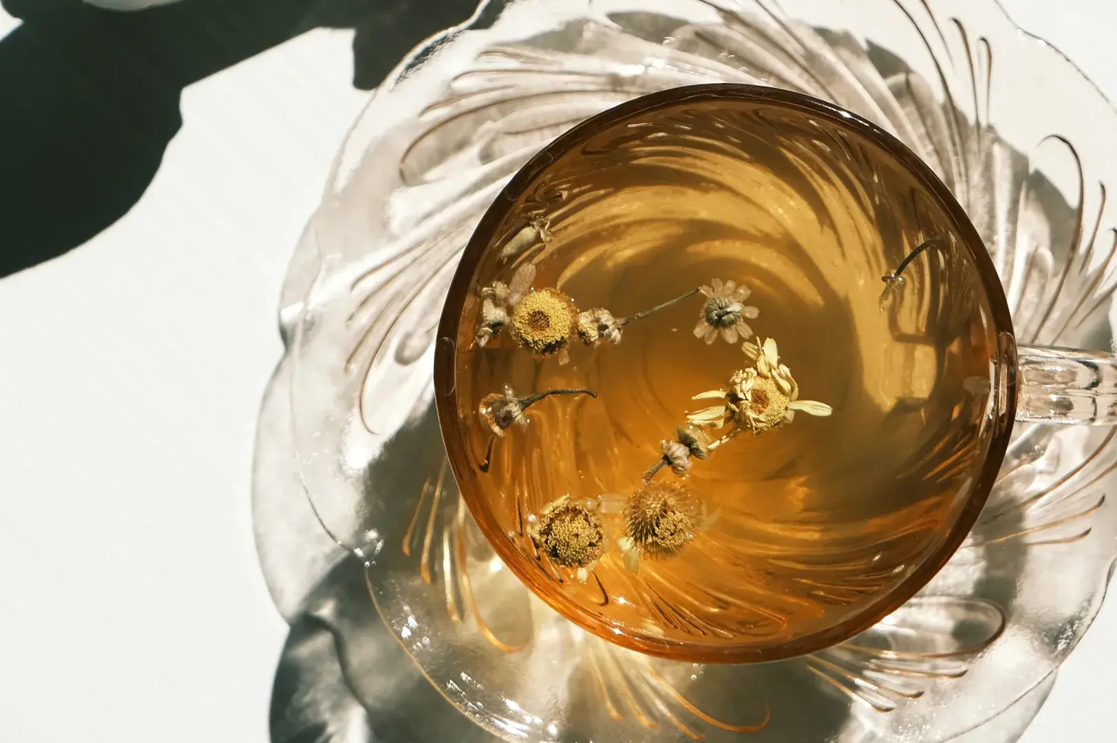 A direct overhead shot highlights a clear amber glass teacup filled with a rich, golden-brown chamomile tea, with several dried chamomile blossoms floating gracefully on the surface. The cup rests on a matching clear textured glass saucer that creates elegant, swirling shadow patterns on the bright white surface below. Strong natural sunlight streams in from the right, casting sharp shadows and illuminating the translucent clarity and warmth of the beverage.