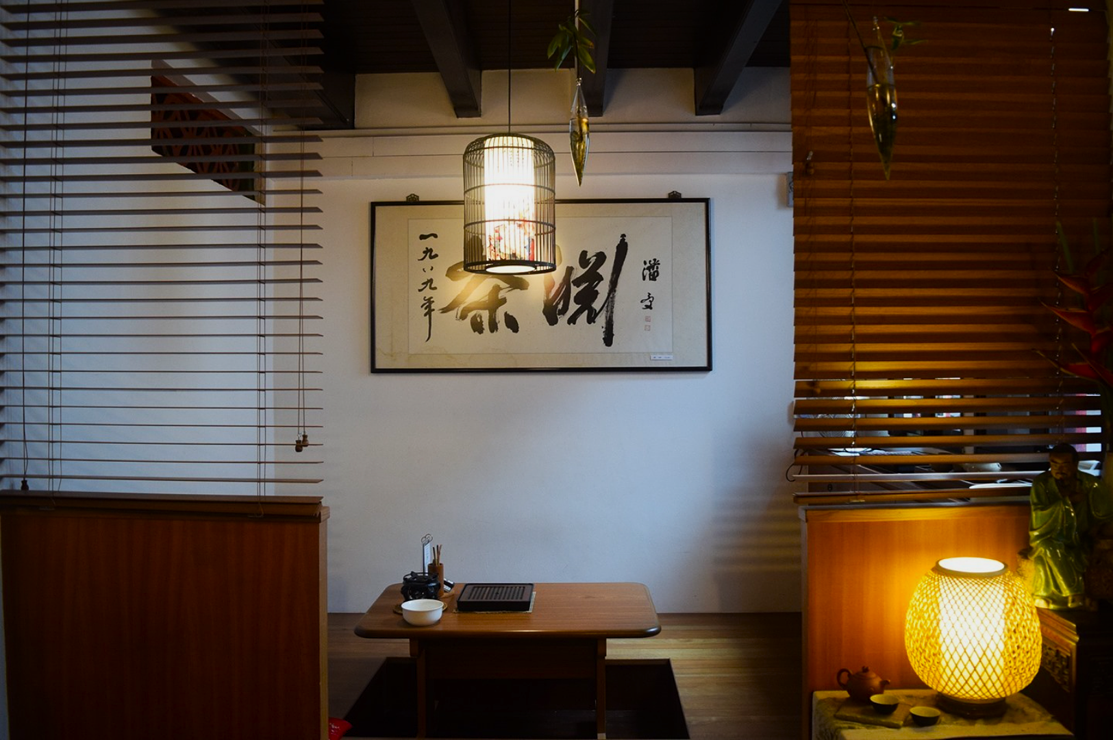 Cozy Japanese-style room with low wooden table, calligraphy art on wall, and warm lantern. Calm and inviting atmosphere with wooden blinds.