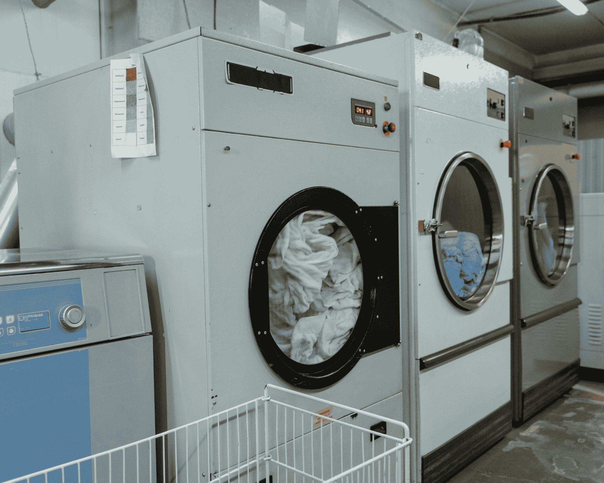 Row of large industrial dryers filled with white linens in a commercial laundry setting.