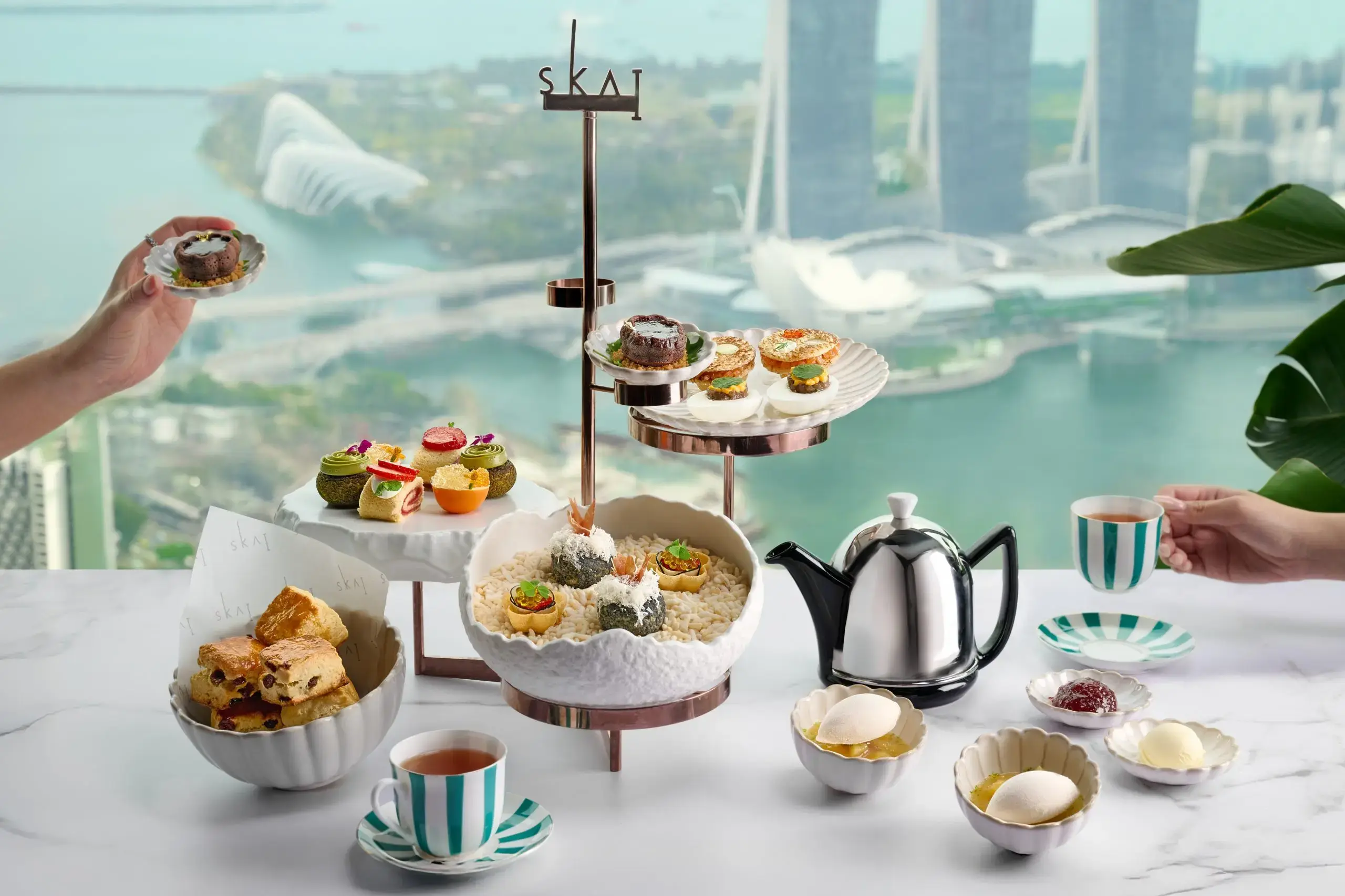 An elegant high tea spread features an assortment of sweet and savory pastries displayed on tiered stands next to a silver teapot and striped teacups. The table is set against a floor-to-ceiling window offering a stunning, elevated view of Singapore's Marina Bay waterfront.