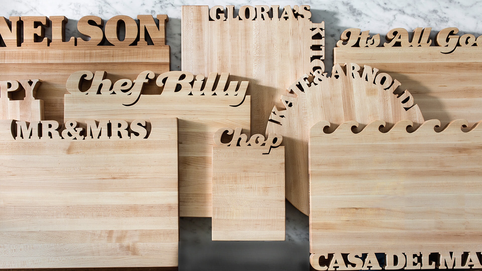 A beautifully arranged collection of personalized wooden kitchen boards and lazy susans against a marble backsplash, showcasing unique designs ideal for home cooks. This display features essential kitchen utensils that make cooking fun and convenient for serving guests and family.