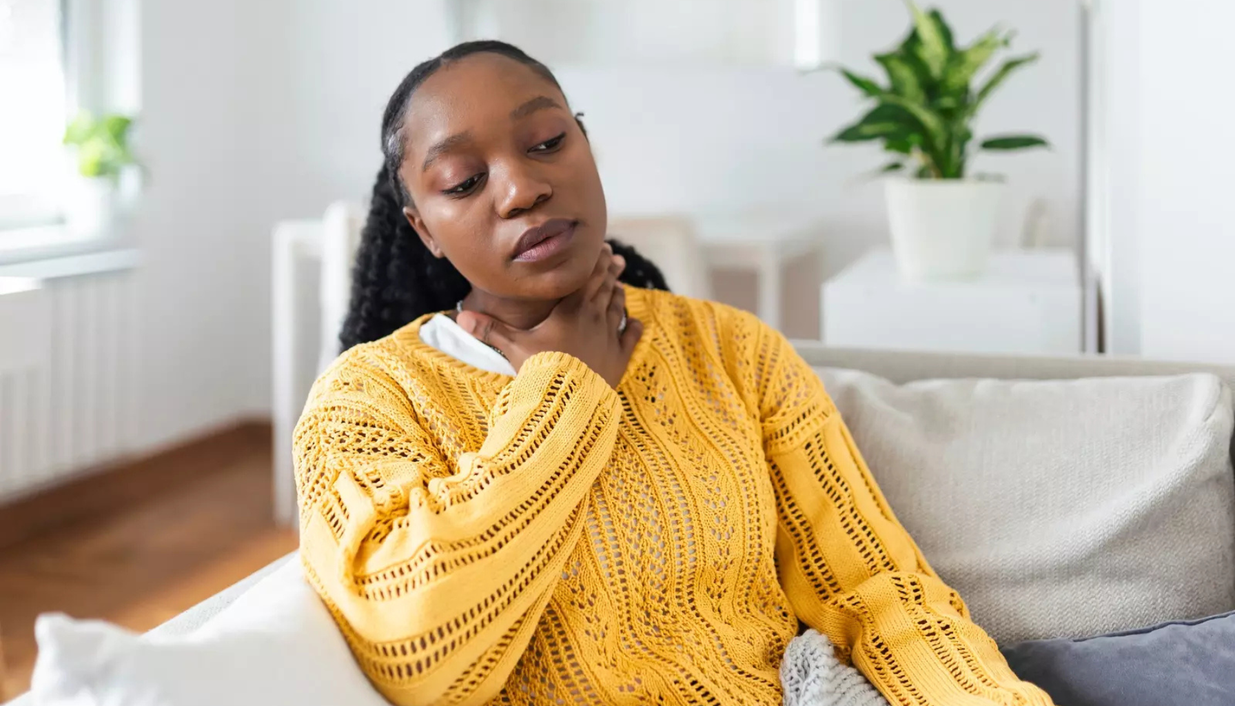 Kvinne sitter i sofaen og holder seg på halsen, muligens på grunn av sår hals eller ubehag.