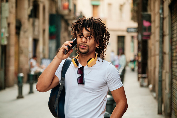 Young man with dark hair and yellow headphones around his neck talking on his cell.