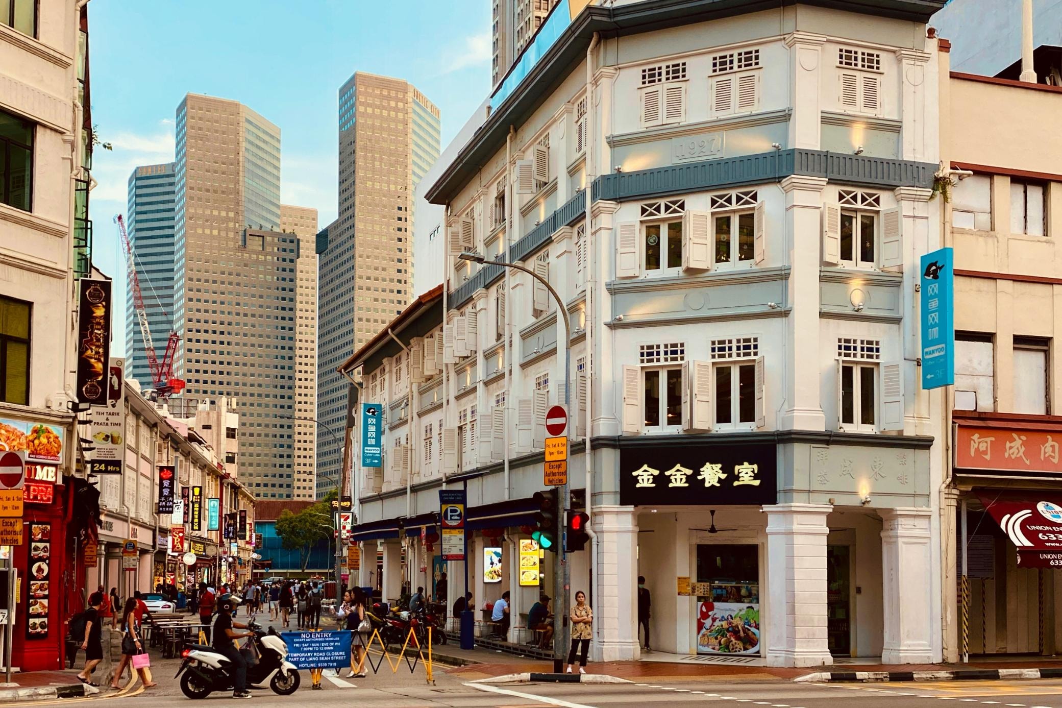 This vibrant street scene features a traditional white corner building with shuttered windows and Chinese signage, contrasting sharply with the towering modern skyscrapers in the background. Pedestrians and a scooter navigate the bustling intersection, which is lined with colorful shops and showcases a distinctive blend of historic charm and contemporary city life.
