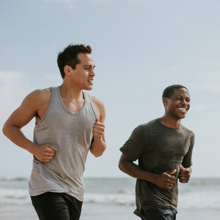 Two young men, one Hispanic and one African, jogging along a sandy beach, displaying vitality and friendship. This image underscores the benefits of supplements and vitamins available at The Good Stuff Health Shop South Africa, part of the TERRA NOVA Collection, where expert advice on health and wellness is readily provided.