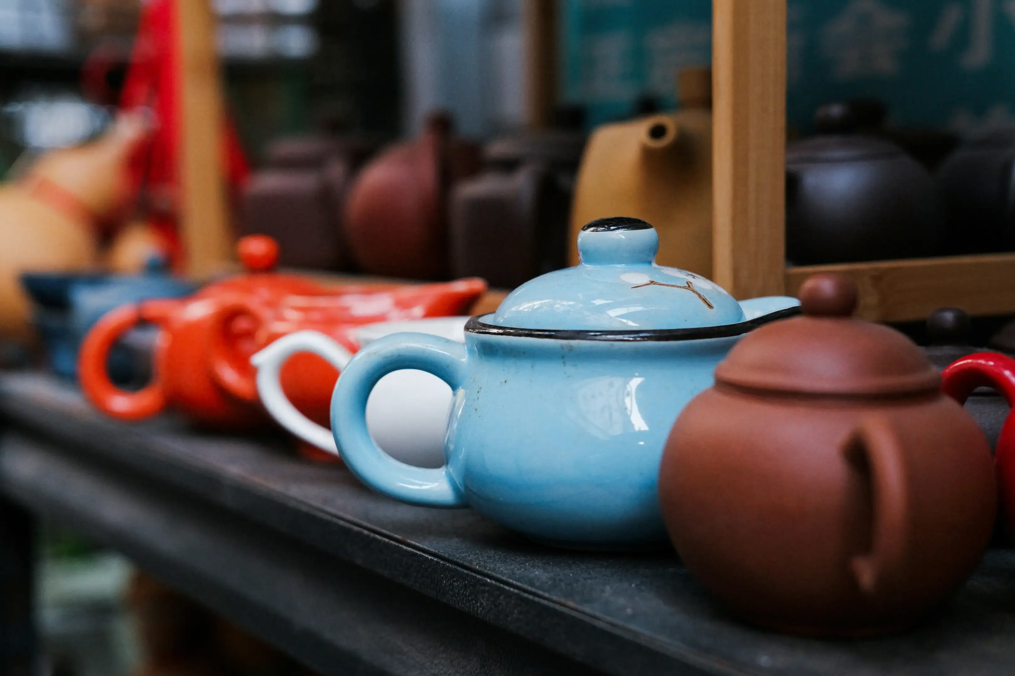 A collection of colorful ceramic teapots, including vibrant red, soft blue, and earthy clay versions, rests on a dark shelf. In the background, additional teaware is displayed within wooden frames, creating a cozy and artisanal atmosphere.