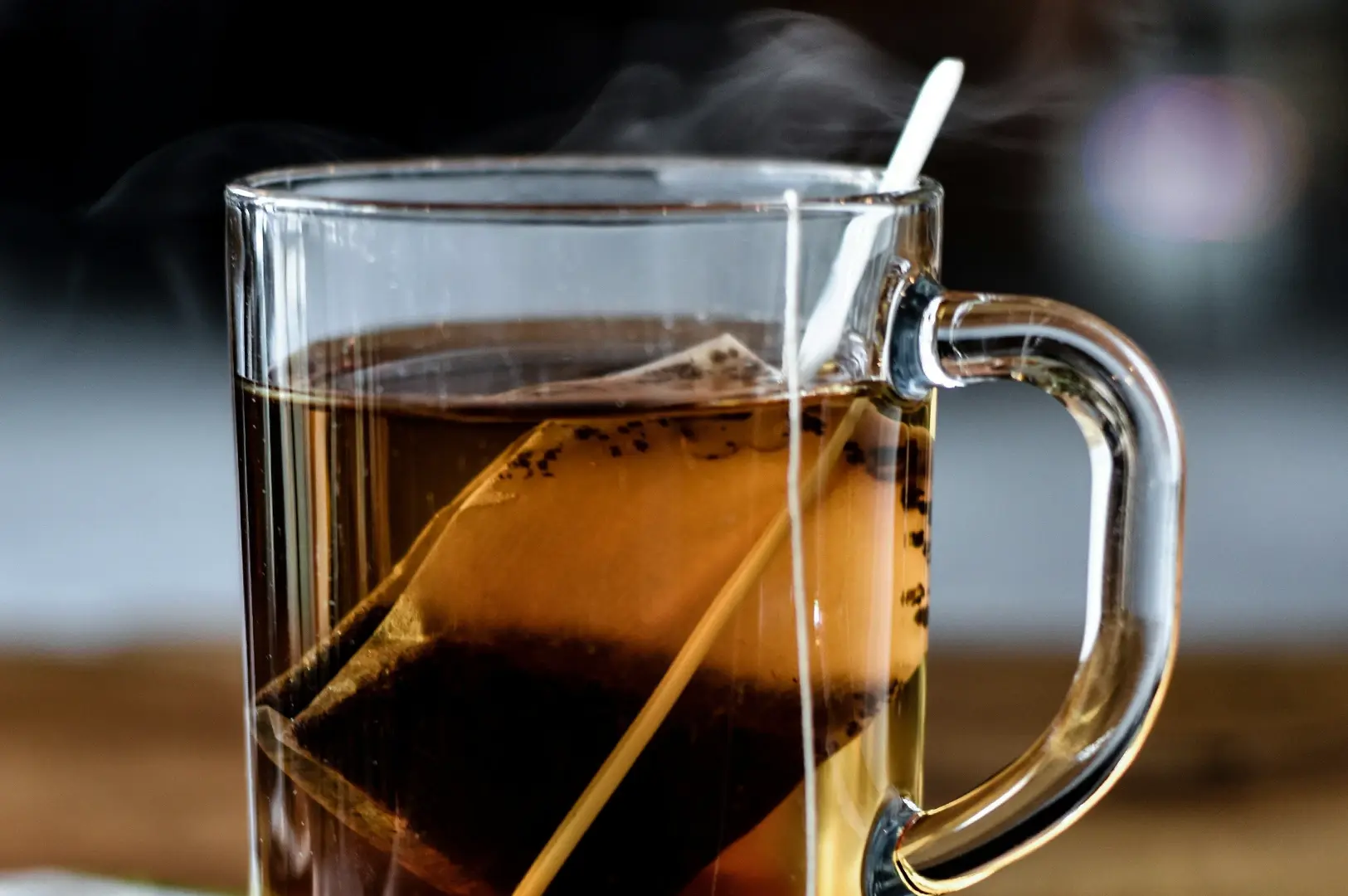 A close-up side view captures a clear glass mug filled with hot, dark tea, with a tea bag still steeping inside. A white tag on a string hangs over the rim of the mug, and delicate wisps of steam rise from the surface of the tea, indicating that the drink is freshly brewed and hot.