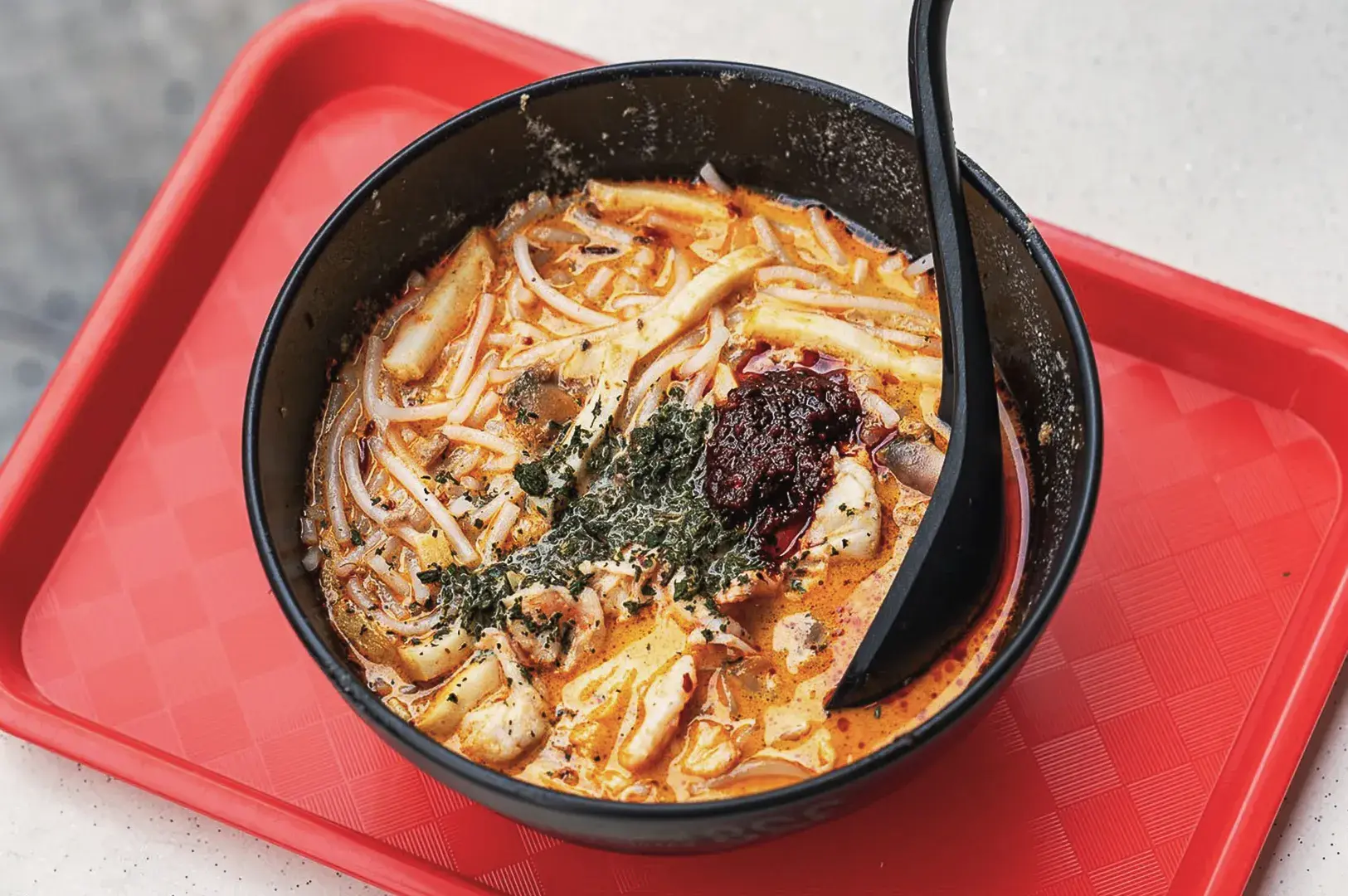 Authentic Singapore laksa noodle soup served in a black bowl with spicy coconut broth, seafood, and sambal chili on a red tray.