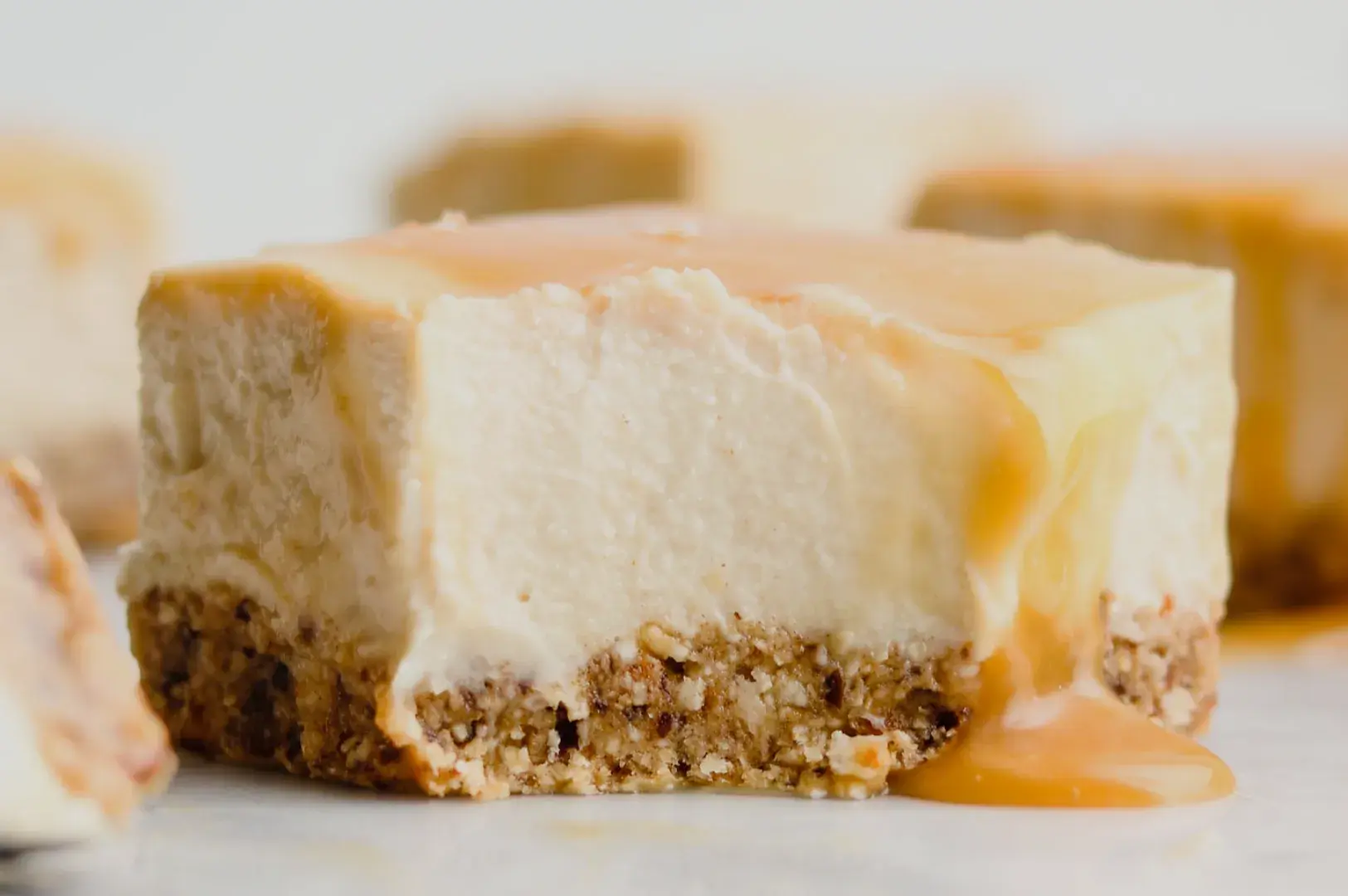 Close-up of a creamy cheesecake bar with a crumbly brown crust, drizzled in glossy caramel; more bars blur in the background, creating a warm, tempting mood.