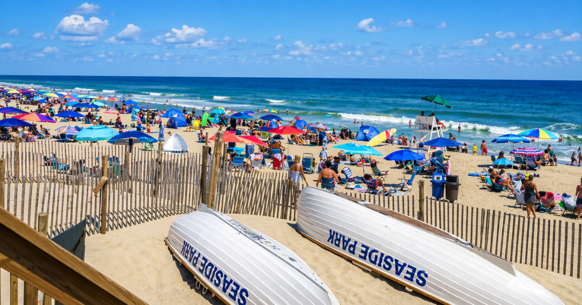 Wide view of Seaside Park beach on a sunny day, featuring colorful umbrellas, beachgoers along the shoreline, gentle ocean waves, and lifeguard stands, with “Seaside Park” boats resting in the sand in the foreground.