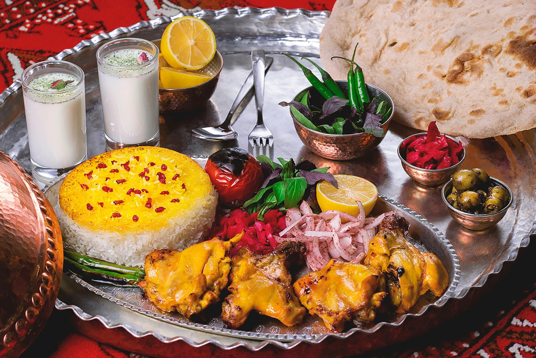  This image showcases a lavish Persian feast served on a large, silver-toned platter, featuring saffron-topped rice and grilled chicken kabobs. The spread is complemented by various side dishes, including charred tomatoes, pickled onions, olives, and fresh herbs, alongside two glasses of doogh and a piece of flatbread.