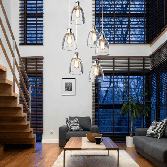 Modern living room with high ceilings, featuring a cluster of glass pendant lights above a wooden coffee table, grey sofas, and large windows with wooden blinds.
