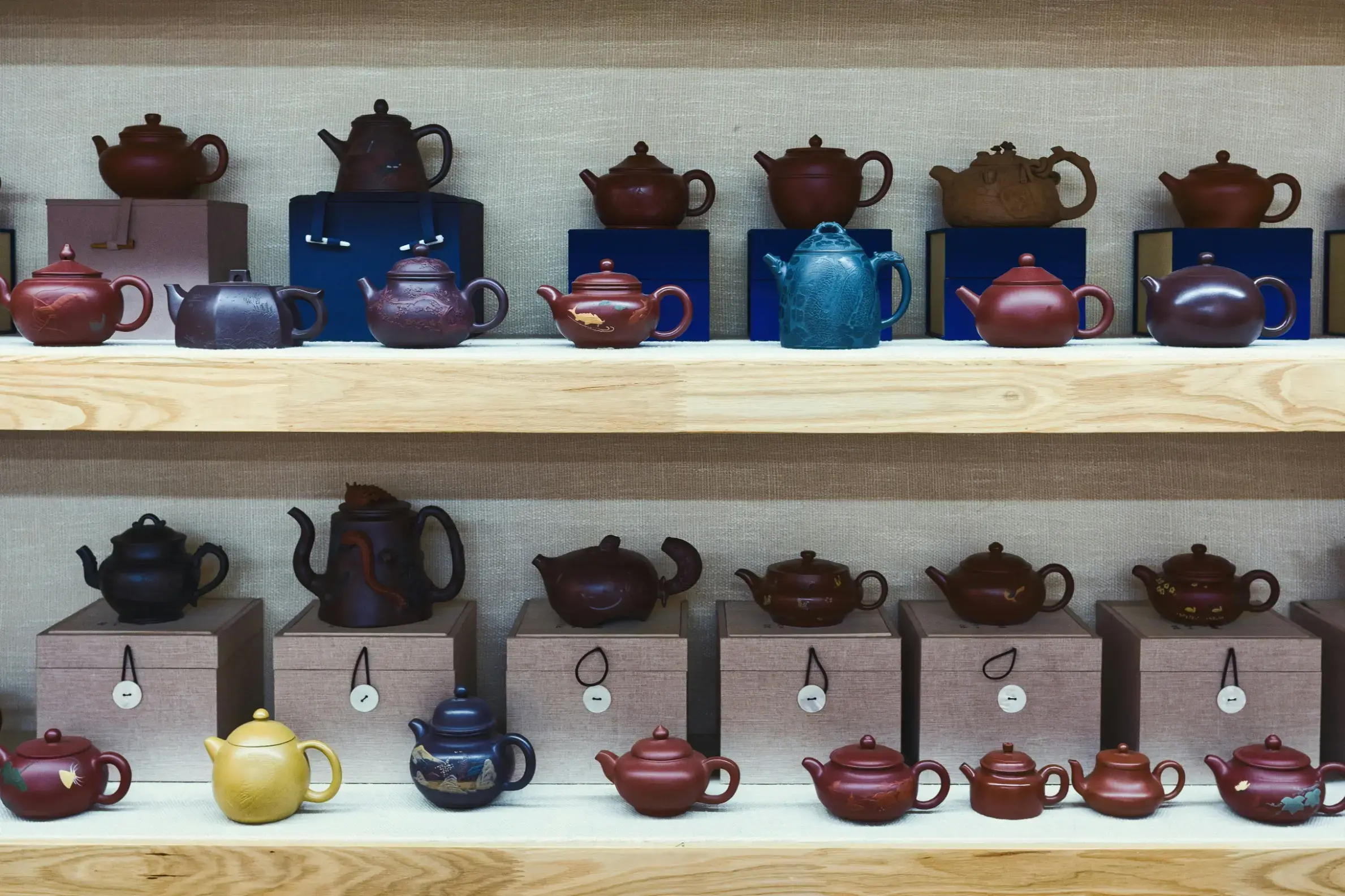 This collection features a variety of traditional clay teapots in diverse earthy tones and textures, neatly arranged across two wooden shelves. Many of the teapots are paired with individual storage boxes, showcasing a mix of simple rounded designs and more intricate, decorative shapes.