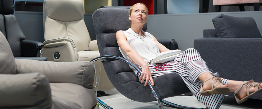 A woman is lying in an rocking armchair with metal armrests, looking up at the ceiling. She is in a showroom surrounded by armchairs.