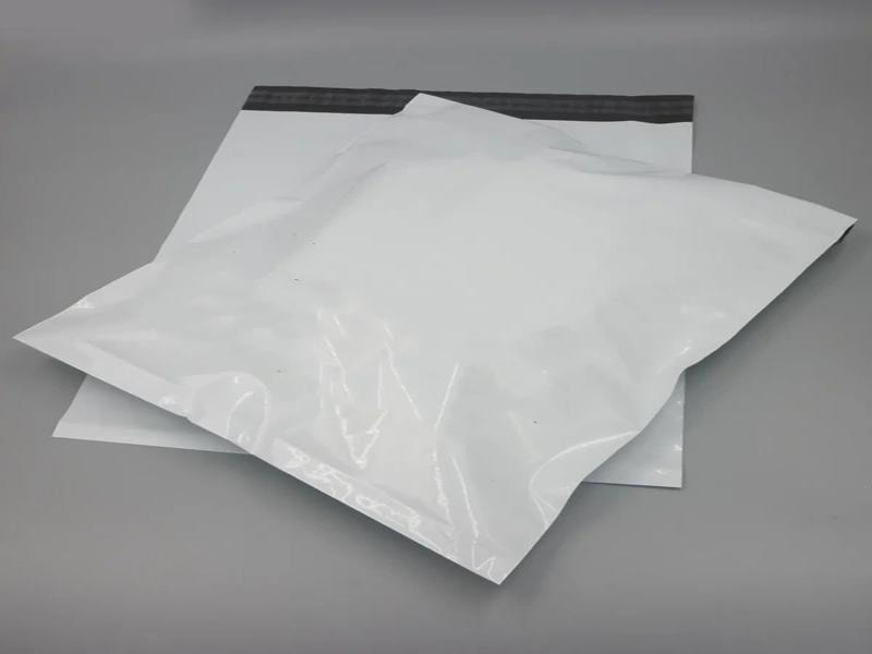 A gray surface. One bag is placed on top of the other, slightly overlapping. The bags are made of a glossy, flexible material, likely polyethylene, commonly used for shipping lightweight items like clothing. The top bag appears to be filled, giving it a slightly puffy shape, while the bottom bag is flat and unopened. Both bags have a black adhesive strip at the opening, which is used to seal them securely. 